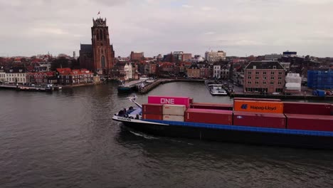 Aerial-Port-Side-View-Of-Sensation-Cargo-Vessel-Going-Past-On-Oude-Maas-With-Our-Lady-of-Dordrecht-In-Background