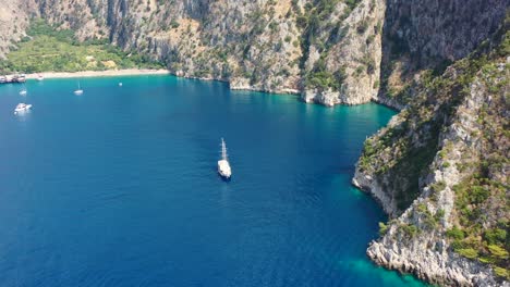 aerial-drone-approaching-a-large-sailboat-docked-in-the-bay-of-Butterfly-Valley-surrounded-by-turquoise-blue-ocean-and-large-mountains-in-Fethiye-Turkey