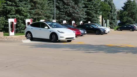Weißes-Tesla-Auto-Verlässt-Die-Super-Ladestation-Auf-Einem-Parkplatz-Für-Ein-Lebensmittelgeschäft-Und-Ein-Restaurant