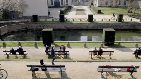 Menschen-Sitzen-Auf-Bänken-Unter-Der-Sonne-Mit-Blick-Auf-Den-Teich-In-Der-Abtei-La-Cambre-In-Ixelles,-Brüssel,-Belgien