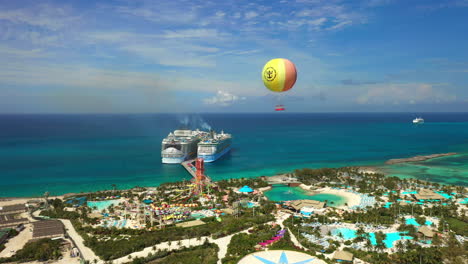 Cinematic-rising-drone-shot-of-CoCoCay-island-with-water-slides-and-a-Royal-Caribbean-cruise-ship-in-the-background,-with-hot-air-ballon
