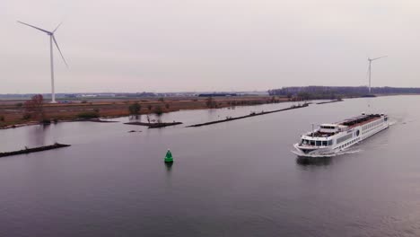 Aerial-View-Of-River-Cruise-Ship-Navigating-Along-Oude-Maas-Going-Past-Still-Wind-Turbines-In-Barendrecht