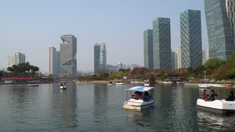 Besucher-Genießen-Die-Tretboote-Auf-Dem-Fluss-Im-Central-Park-In-Incheon,-Südkorea,-Mit-Der-Skyline-Der-Stadt-Im-Hintergrund