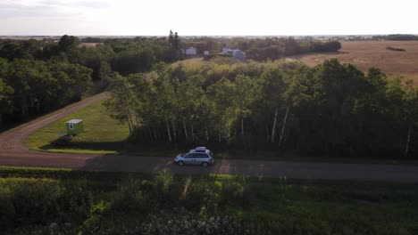 4k-aerial-shot-of-silver-minivan-driving-slowly-along-dirt-road-next-to-trees-during-sunset-in-rural-countryside-setting