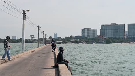 Fisherman-sitting-on-the-pier-smoking-while-holding-his-rod-and-then-another-one-approaches-to-thrown-some-chumming-material-in-the-water,-Pattaya-Fishing-Dock,-Chonburi,-Thailand