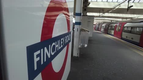 Close-Up-Side-View-Of-Finchley-Road-Roundel-Station-Sign-With-Northbound-Jubilee-Train-Arriving-At-Platform-On-12-May-2022