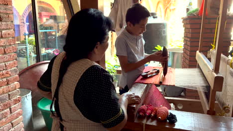 Inside-Traditional-Artisan-Rugs-Workshop-Manufacture,-Woman-Teaching-Watching-Kid-Practicing-Manipulating-Weaving-Braiding-Machine,-Apprentice-Craftsman-Worker-Working,-Artisan-Handloom-Craftwork