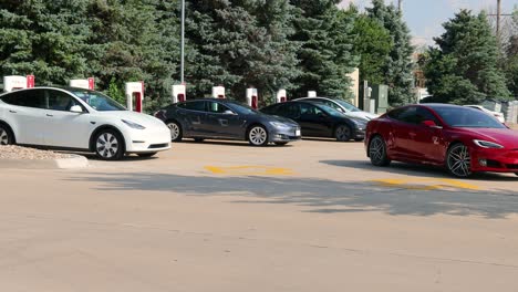 Coche-Tesla-Rojo-Saliendo-De-Una-De-Las-Estaciones-De-Supercarga-En-Un-Estacionamiento-Para-Una-Tienda-De-Comestibles-Y-Un-Restaurante