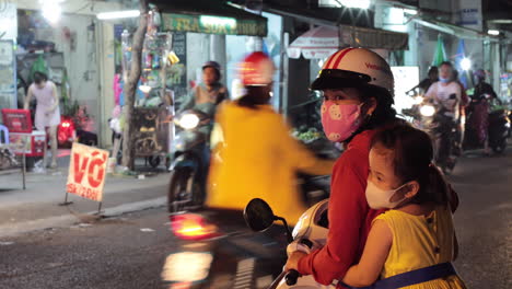 A-mother-and-young-daughter-try-to-pull-in-to-the-steady-stream-of-motor-scooter-traffic-at-night