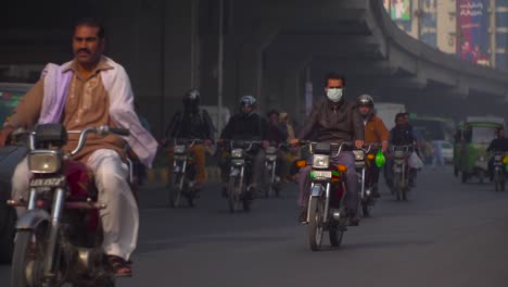 Traffic-close-up-at-the-road,-Man-wore-mask-on-the-bike-instead-of-a-helmet,-passing-motor-bikes-and-cars-under-the-bridge,-Buses,-rikshaw`s,-billboards-and-buildings-in-the-background