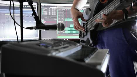 Musician-playing-bass-guitar-in-a-home-studio-with-PC-screens-displaying-software