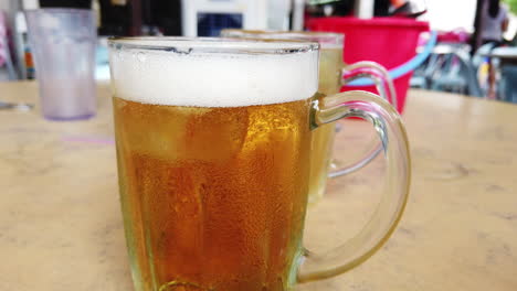 Footage-of-2-Asian-beer-mug-placed-on-the-table-being-poured-with-beer-with-other-table-and-people-seen-in-the-background