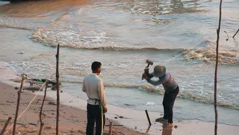Medium-Exterior-Shot-of-Man-Hammering-Post-Into-the-Sand-With-a-Heavy-Hammer-and-his-Friend-on-the-Shore-With-Waves-in-the-Water-Behind-Him-in-Lake-in-the-Daytime