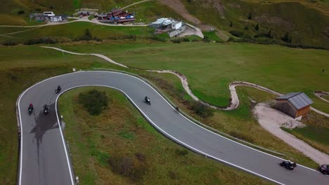 Group-of-bikers-traverses-a-winding-road-along-a-green-pasture-covered-mountain-pass-in-the-Dolomite-Italian-Alps,-Drone-dolly-out-reveal-shot