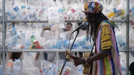 Street-artist-singing-in-a-park-performance,-Barcelona-in-front-of-plastic-bottle-wall-artwork-piece-against-plastic-pollution