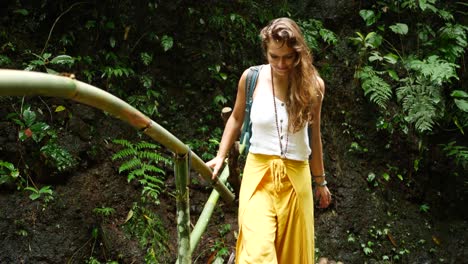 A-young-attractive-woman-walks-over-a-small-bamboo-bridge-over-a-small-stream-of-water-being-happy-with-a-smile-in-a-beautiful-and-lush-jungle-in-Bali