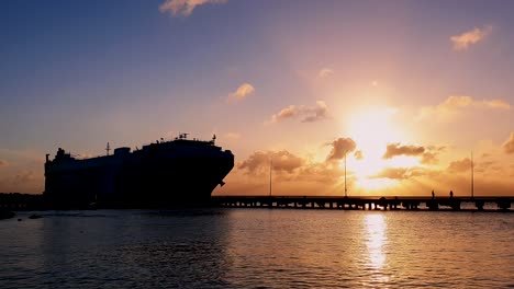 Silhouette-of-cargo-ship-for-international-trade-approaching-for-docking-in-harbor-during-beautiful-sunset,-wide-shot