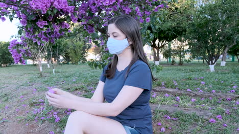 Caucasian-woman-with-face-mask-sitting-in-park-collecting-pink-flower-from-spring-tree