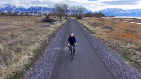 Mature,-older-man-bicycling-along-a-nature-path-with-scenic,-snowcapped-mountains-in-the-distance---leading-aerial-view