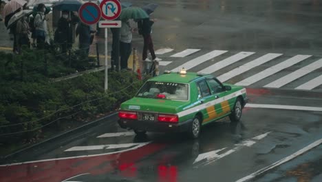 Taxi,-Das-An-Der-Shibuya-Kreuzung-Nach-Links-Abbiegt,-Mit-Menschen,-Die-An-Einem-Regnerischen-Tag-In-Tokio,-Japan,-Einen-Regenschirm-Halten