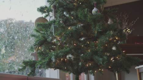 Auf-Der-Anderen-Seite-Ist-Ein-Mit-Lichtern-Und-Dekorationen-Bedeckter-Weihnachtsbaum-Neben-Einem-Fenster-Zu-Sehen,-Durch-Den-Schnee-Fällt