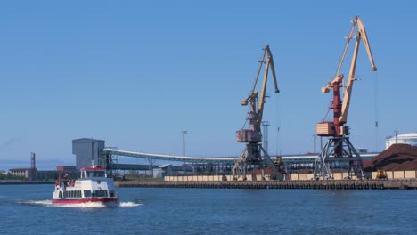 Pleasure-boat-Hercogs-JÄ“kabs-with-people-on-the-deck-moving-at-Venta-river,-port-cranes-and-harbor-warehouses-in-background,-sunny-summer-day,-Port-of-Ventspils,-wide-shot-from-a-distance