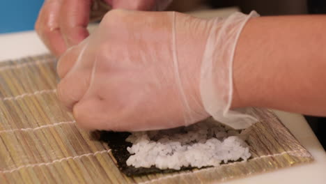 Hände-Verteilen-Schnell-Sushi-Reis-Auf-Nori-Wrap-Mit-Bambusmatte-Darunter---Nahaufnahme