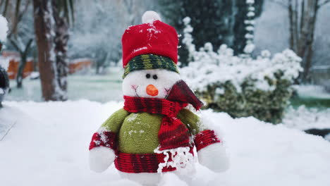 A-snowman-sof-toy-is-sitting-on-a-table-in-a-garden-as-snow-falls-around-him