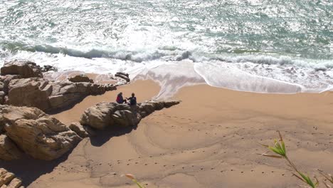 Aerial-view-of-couple-sitting-on-rocks-watching-ocean-waves-on-the-beach