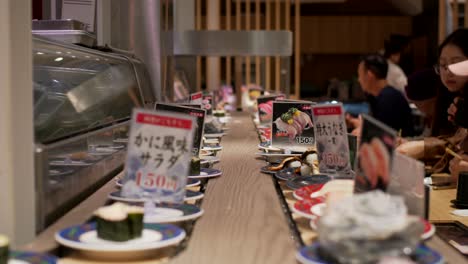 Plates-Of-Sushi-On-The-Conveyor-Belt-Moving-In-Front-Of-Customers-Sitting-And-Eating-Inside-A-Kaitenzushi-Restaurant---Kaiten-sushi-Conveyor-Belt-Sushi-In-Kyoto,-Japan