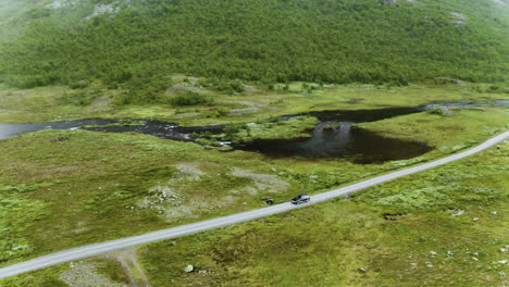 Car-Driving-On-The-Long-And-Winding-Road-Along-The-Lush-Field-And-Calm-Lake-In-Hydalen,-A-Hidden-Gem-In-Hemsedal,-Norway-On-A-Misty-Morning---aerial-drone