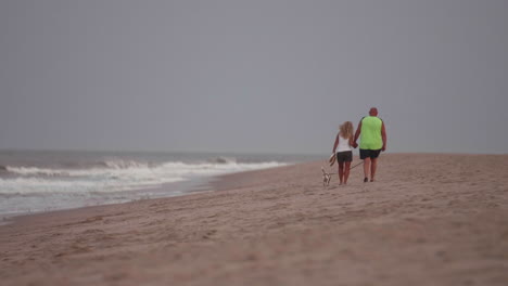 Middle-aged-couple-holding-hands-and-walking-dog-on-beach-at-sunset,-Slow-Motion
