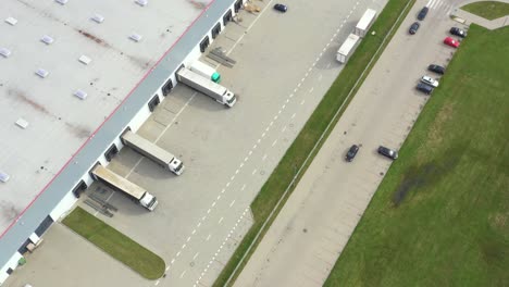 Aerial-following-shot-of-semi-trailer-trucks-travelling-along-a-warehouse-on-the-parking-lot-in-the-logistics-park-with-loading-hub