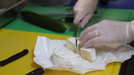 Die-Hand-Des-Kochs-Mit-Handschuh-Schneidet-Runden-Brie-Käse-Mit-Einem-Messer-Auf-Einem-Schneidebrett
