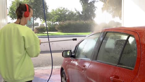woman-cleaning-roof-of-red-car-with-pressure-hose-while-listening-to-music-with-mask-on