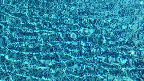 Crystal-clear-water-in-a-swimming-pool-with-sunlight-casting-shadows-in-the-water-and-on-the-tiled-floor---textured-background