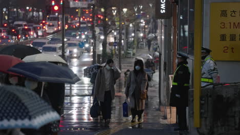 Zwei-Sicherheitskräfte-Beobachten-Menschen,-Die-In-Einer-Regnerischen-Nacht-Während-Einer-Pandemie-In-Tokio,-Japan,-Auf-Der-Straße-Gehen