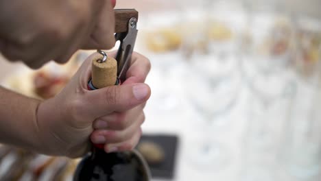 Kaukasische-Hand-öffnet-Eine-Flasche-Rotwein-Mit-Flaschenöffner-Mit-Echtem-Kork-Und-Das-Catering-Im-Hintergrund