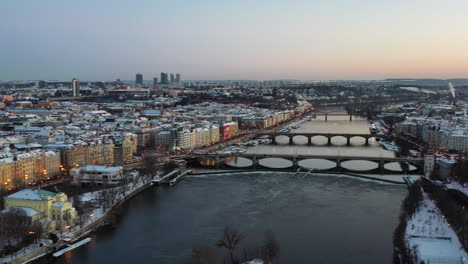 Aerial-view-of-Prague,-Czech-Republic-at-winter-twilight-bridges-on-Vltava-river-and-snow-capped-buildings,-panoramic-drone-shot