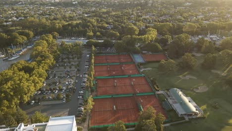 Aerial-cinematic-shot-of-a-private-sport-club-with-tennis-courts,-a-golf-field-and-a-parking-lot-at-golden-hour