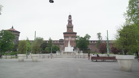 Frontal-view-of-"Castello-Sforzesco",-with-fountain,-people-walking-and-motorbikes-passing-by