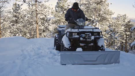 A-Caucasian-male-sits-down-on-an-ATV-or-4-wheeler-with-a-plow-and-drives-forward-to-clear-snow-from-a-mountain-driveway