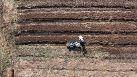 AERIAL---Man-working-a-field-with-a-rototiller,-agriculture,-wide-shot-zoom-in