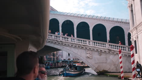POV-Cámara-Lenta-Tomada-Desde-Un-Barco-Que-Muestra-El-Famoso-Puente-De-Rialto-Con-Turistas-Durante-Un-Día-Soleado-En-Venecia,-Italia