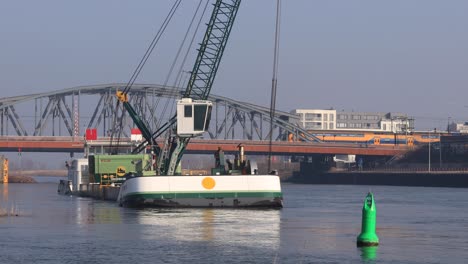 Ship-with-large-crane-dredging-the-river-IJssel-cargo-ship-corridor-leading-underneath-the-draw-bridge-behind-with-a-yellow-blue-train-of-the-Dutch-national-railway-passing-by-in-the-background