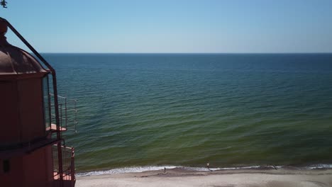 Beautiful-aerial-view-of-white-painted-steel-lighthouse-with-red-top-located-in-Pape,-Latvia-at-Baltic-sea-coastline-in-sunny-summer-day,-sea-in-background,-wide-angle-drone-shot-moving-forward-close