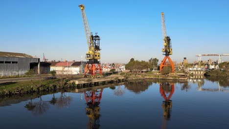 Low-backward-aerial-of-port-cranes-by-still-water-in-Buenos-Aires