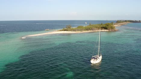 Sailboat-Floating-In-Shallow-Sea-Water-With-Beautiful-Coral-Reef-At-The-Bottom-And-Tropical-Paradise-Island-In-Background