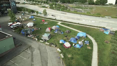 Un-Pequeño-Campamento-Para-Personas-Sin-Hogar-En-Un-Espacio-Verde-Urbano-A-Lo-Largo-De-Una-Carretera,-Aéreo