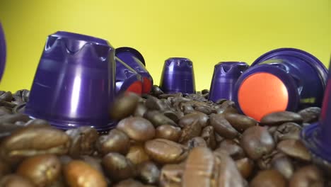 Close-up-shot-of-falling-roasted-coffee-beans-between-coffee-capsule-in-front-of-yellow-background